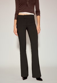 Crne hlače trapezastog kroja s glatkom teksturom, jednostavnim pojasom i prilagođenim krojem kroz bedro, uparene s pripijenim smeđim topom.