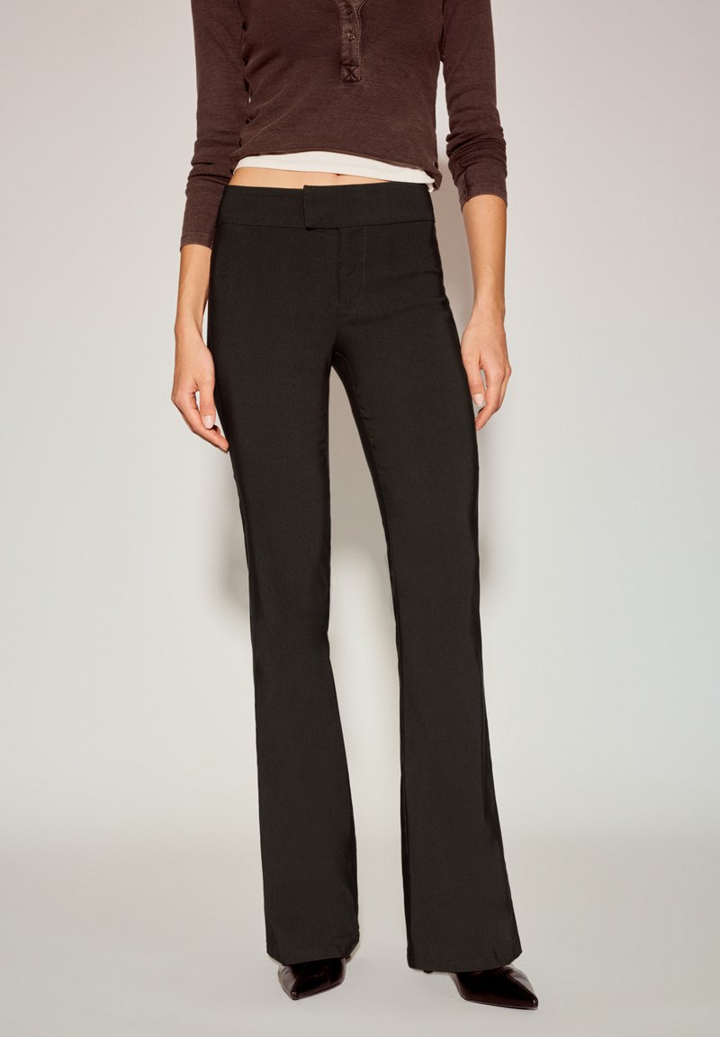 Crne hlače trapezastog kroja s glatkom teksturom, jednostavnim pojasom i prilagođenim krojem kroz bedro, uparene s pripijenim smeđim topom.