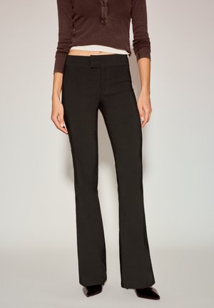 Pantalon noir évasé avec une texture lisse, une ceinture simple et une coupe ajustée au niveau de la cuisse, associé à un haut marron moulant.