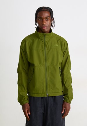 Man standing facing forward wearing a zipped green North Face jacket and loose black pants against a plain light background.