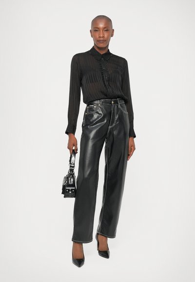 Schwarze, durchsichtige Bluse mit Knopfverschluss und Kragen über hochtaillierten schwarzen Kunstlederhosen, kombiniert mit schwarzen spitz zulaufenden Heels und einer kleinen Tasche.