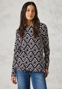 Langärmliges Oberteil in Schwarz-Weiß mit einem Diamantmuster, Saum mit Kordelzug und weichem Stoff. Kombiniert mit blauen Jeans.