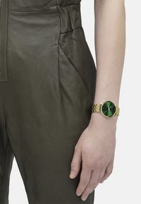 Goldene Armbanduhr mit einem runden grünen Ziffernblatt und dünnen Stundenmarkierungen, getragen an einem hellen Hautton-arm, vor einem olivgrünen Leder-Overall.