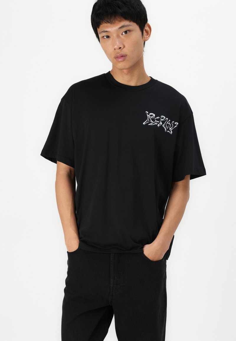 Camiseta negra de manga corta hecha de algodón con un corte relajado, que presenta texto gráfico blanco en el lado izquierdo del pecho y una textura suave.