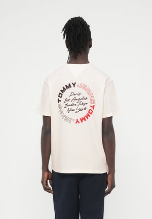 Osoba s tmavou pletí a dredy nosí béžové tričko s kruhovým nápisem "Tommy Jeans" a názvy měst na zádech, spojené s tmavými kalhotami.