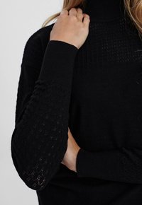 Haut du torse d'une femme avec une main touchant le cou, portant un pull en tricot noir avec des manches texturées et un col montant côtelé.