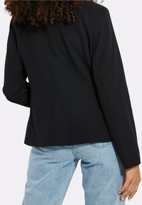 Blazer noir ajusté, à manches longues et en tissu lisse. Le dos présente un détail de couture. Associé à un jean en denim bleu clair.