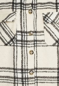 Krämfärgad rutig skjorta med svarta rutor, med två bröstfickor, knäppning med knappar och strukturerad tyg. Runda bruna knappar.