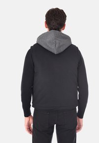 Gilet nero con cappuccio grigio testurizzato, caratterizzato da una texture a coste e un design aderente. La vista posteriore mostra hardware minimale e linee pulite.