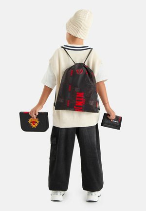 Bambino con cappello di lana color crema e gilet, pantaloni larghi neri, che porta uno zaino con coulisse nero e rosso e tiene in entrambe le mani dei portafogli neri.