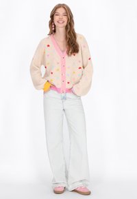 Cardigan tricotat bej cu modele colorate de inimi, mansete și margini roz, asortat cu blugi deschiși la culoare și evazați și pantofi roz.