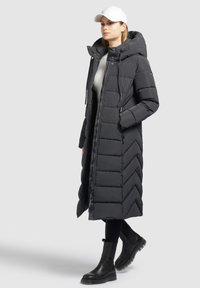 Cappotto imbottito lungo di un grigio scuro, con trapuntatura a zig zag, grande cappuccio, zip anteriore e tasche laterali. Indossato con leggings neri e stivali neri voluminosi.