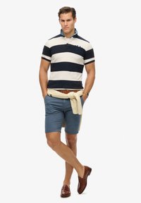 Polo a righe in blu navy e crema, con colletto e taschino sul petto. Pantaloni corti azzurri. Cintura marrone e mocassini. Maglione annodato in vita.