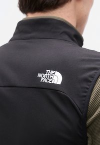Άτομο που φοράει μαύρο γιλέκο North Face πάνω από μια ελιάς πράσινου χρώματος ανάγλυφη μπλούζα με μακριά μανίκια, φαίνεται από πίσω, εμφανίζοντας το λογότυπο στο γιλέκο.