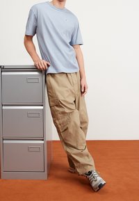 Světle modré bavlněné tričko s malým logem, spárované s béžovými cargo kalhotami s bočními kapsami a texturovanou látkou, stojící poblíž skříně na dokumenty.