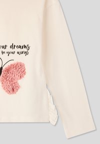 Długo rękawowy kremowy top z różowym teksturowanym wzorem motyla i czarnym nadrukowanym tekstem. Po boku znajduje się falbanka wzdłuż dołu.