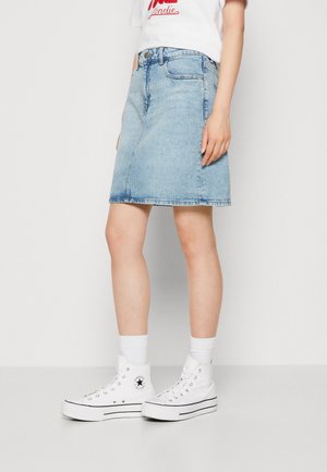 Person iført lysblåt denimskørt, hvide høje sneakers med sort stribe og logo, hvide sokker og en del af en hvid t-shirt med rød tekst.