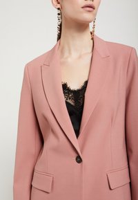 Ružový priliehavý blejzer s klopnými límčekmi, zapínaním na jeden gombík a prednými vreckami. Pod ním je viditeľný čierny top s čipkovým lemovaním.