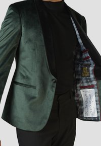 Grüner Samtblazer mit schwarzem Satinrevers, Einzelknopfverschluss, seitlicher Tasche und kariertem Futter mit einer Anleitung zum Binden einer Fliege.