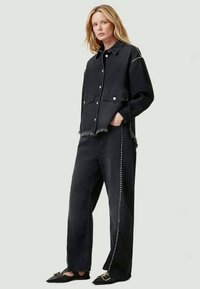 Chaqueta de denim negra con botones y dobladillo con flecos, combinada con pantalones a juego que tienen remaches laterales. La modelo lleva zapatos planos negros.