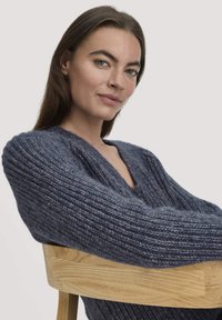 Dunkelblauer V-Ausschnitt-Strickpullover mit gerippter Textur und langen, lockeren Ärmeln. Das Model sitzt mit verschränkten Armen auf einem Holzstuhl.
