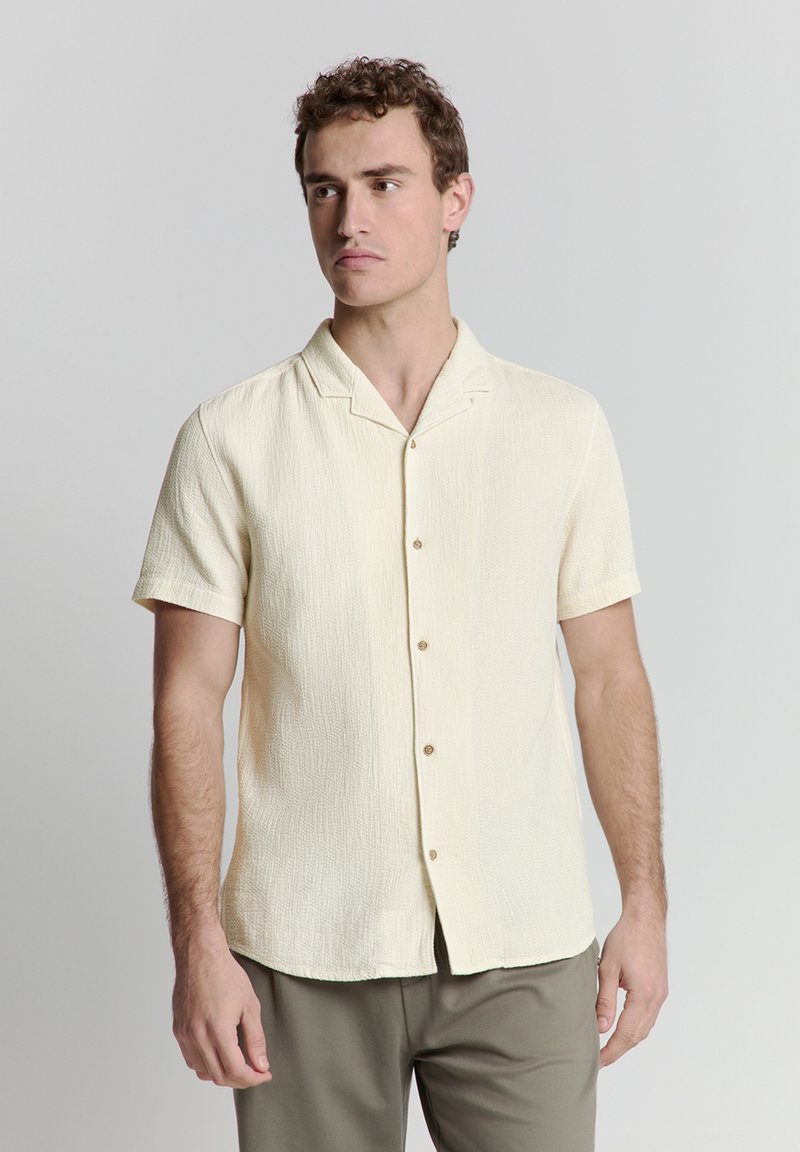 Korte mouw crèmekleurig shirt van gestructureerde stof, voorzien van een ingesneden kraag en bruine knopen aan de voorkant, gedragen met lichte broek.