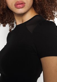 Femme aux cheveux bouclés portant un haut noir à manches courtes avec un détail en mesh transparent sur l'épaule, lèvres légèrement entrouvertes, et légères taches de rousseur sur la peau.