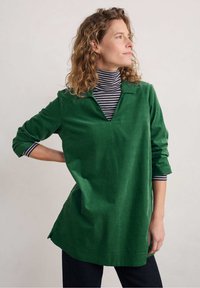 Tunic in velluto a coste verde con scollo a V, dotata di maniche lunghe arrotolate, indossata su un dolcevita a righe bianche e nere. Fit casual e morbido.