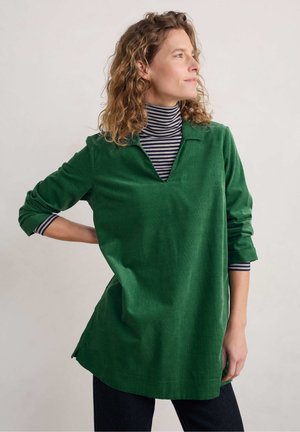 Grøn kordfløjs tunika med V-udskæring, med lange ærmer oprevet, over en sort og hvid stribet turtleneck. Afslappet, løs pasform.