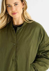 Olive green bomber jacket with a smooth texture, snap buttons, ribbed collar, and slightly oversized fit. Black shirt visible underneath.