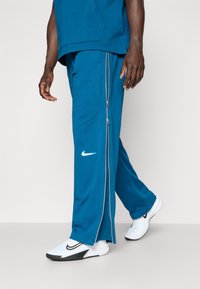 Pantalons de sport teal Nike avec deux zips sur les côtés, une taille élastique et un logo blanc. Accompagnés de chaussures de sport blanches avec des accents noirs.