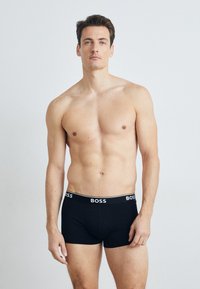 Musta boxeri, joissa on valkoinen "BOSS" -logo vyötäröllä, pehmeää kangasta, istuva malli, keskipituiset lahkeet. Malli poseeraa yksinkertaista taustaa vasten.