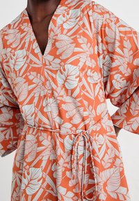 Römdress med ett livligt orange tyg med ett blommönster i vitt, framhävd av breda ärmar och ett knytbälte i midjan.