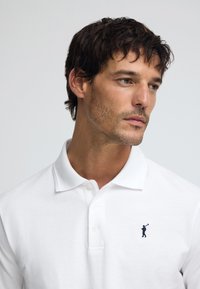 Wit poloshirt met een drukknoopsluiting van drie knopen, voorzien van een klein marineblauw logo van een golfer geborduurd op de linkerborst. Soepele, gestructureerde stof.