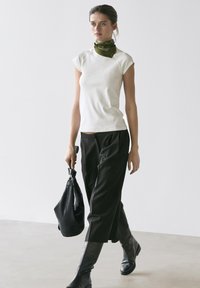 Vrouw die binnen loopt, gekleed in een wit topje met kapmouwen, zwarte wijde pijpen broek, zwarte laarzen, olijfgroene nek sjaal en draagt een zwarte leren handtas.