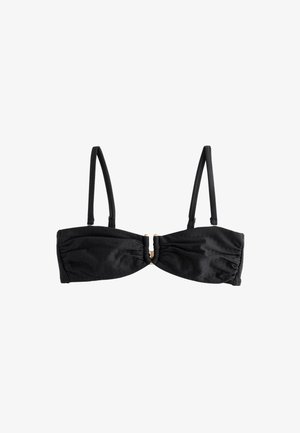 Kobieta z kręconymi włosami ubrana w czarny top bikini widziana od tyłu na jasnoniebieskim tle.