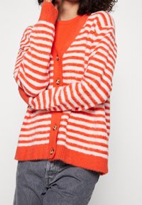 Pruhovaný kardigan oranžové a bílé barvy s měkkou texturou, véčkovým výstřihem a dřevěnými knoflíky vpředu, doplněný jednobarevným oranžovým topem a šedými kalhotami.