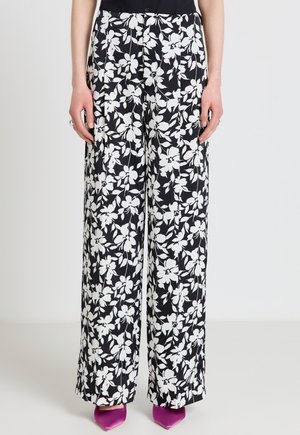 Schwarze, hoch taillierte, weit geschnittene Hose mit weißem Blumenmuster, kombiniert mit einem schwarzen Oberteil und violetten spitzen Absätzen.