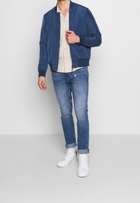 Navy bomberjacka med ribbstickad krage, framsida med dragkedja och sidofickor, kombinerad med en ljusfärgad skjorta med knappar och blå jeans. Vita sneakers.