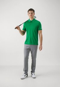 Grön pikétröja med treknappsknäppning, kombinerad med ljusgrå byxor och vita skor, hållande i en golfklubba.