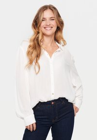 Camicia bianca con bottoni, dal taglio rilassato, con maniche lunghe e colletto. Abbinata a jeans blu scuro con cuciture arancioni.
