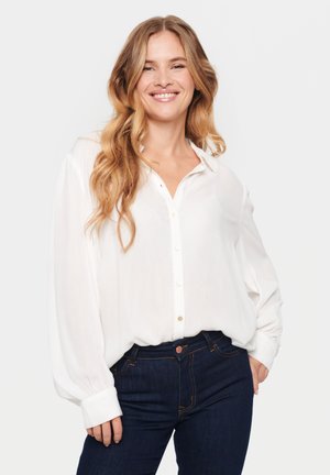 White, button-up blouse with a relaxed fit, long sleeves, and a collar. Paired with dark blue jeans featuring orange stitching.