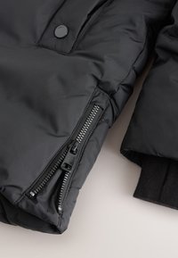 Veste matelassée noire à l'extérieur lisse, avec une fermeture avant à glissière et boutons-pression, des poignets côtelés et une texture matelassée.
