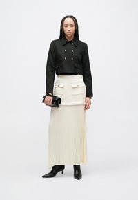 Orezaná čierna bunda so zlatými gombíkmi, kombinovaná s krémovou plisovanou sukňou s vreckami, doplnená čiernymi členkovými čižmami a kabelkou do ruky.