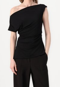 Czarny top odkrywający ramiona, wykonany z miękkiego materiału, z bocznymi marszczeniami, z krótkimi rękawami i luźnym krojem, zestawiony z dopasowanymi czarnymi spodniami.