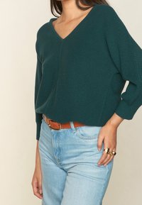 Sweater de punto en teal oscuro con cuello en v y mangas tres cuartos, combinado con jeans de tiro alto en azul claro y un cinturón marrón. Anillos de oro en los dedos.