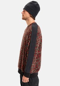 Il maglione presenta un corpo in pile a motivi rossi e verdi, con accenti neri sulle spalle e polsini a coste. Abbinato a un berretto scuro.
