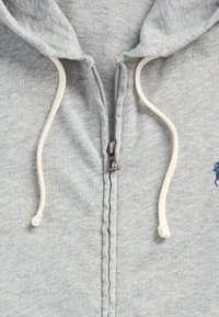 Felpa grigia con chiusura a zip, cordini bianchi e logo ricamato blu. Il tessuto sembra morbido, con dettagli di cucitura visibili.