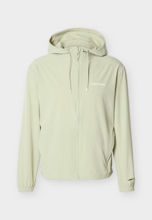 Világos bézs színű Calvin Klein kapucnis dzseki teljes elülső cipzárral, húzózsinórokkal, elasztikus mandzsettákkal és diszkrét logóval a mellkasán.