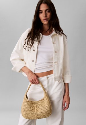 Γυναίκα με μακριά σκούρα μαλλιά, φορώντας λευκό σακάκι, λευκό τοπ και λευκό παντελόνι, κρατώντας μια πλεκτή μπεζ τσάντα με το λογότυπο "CK".